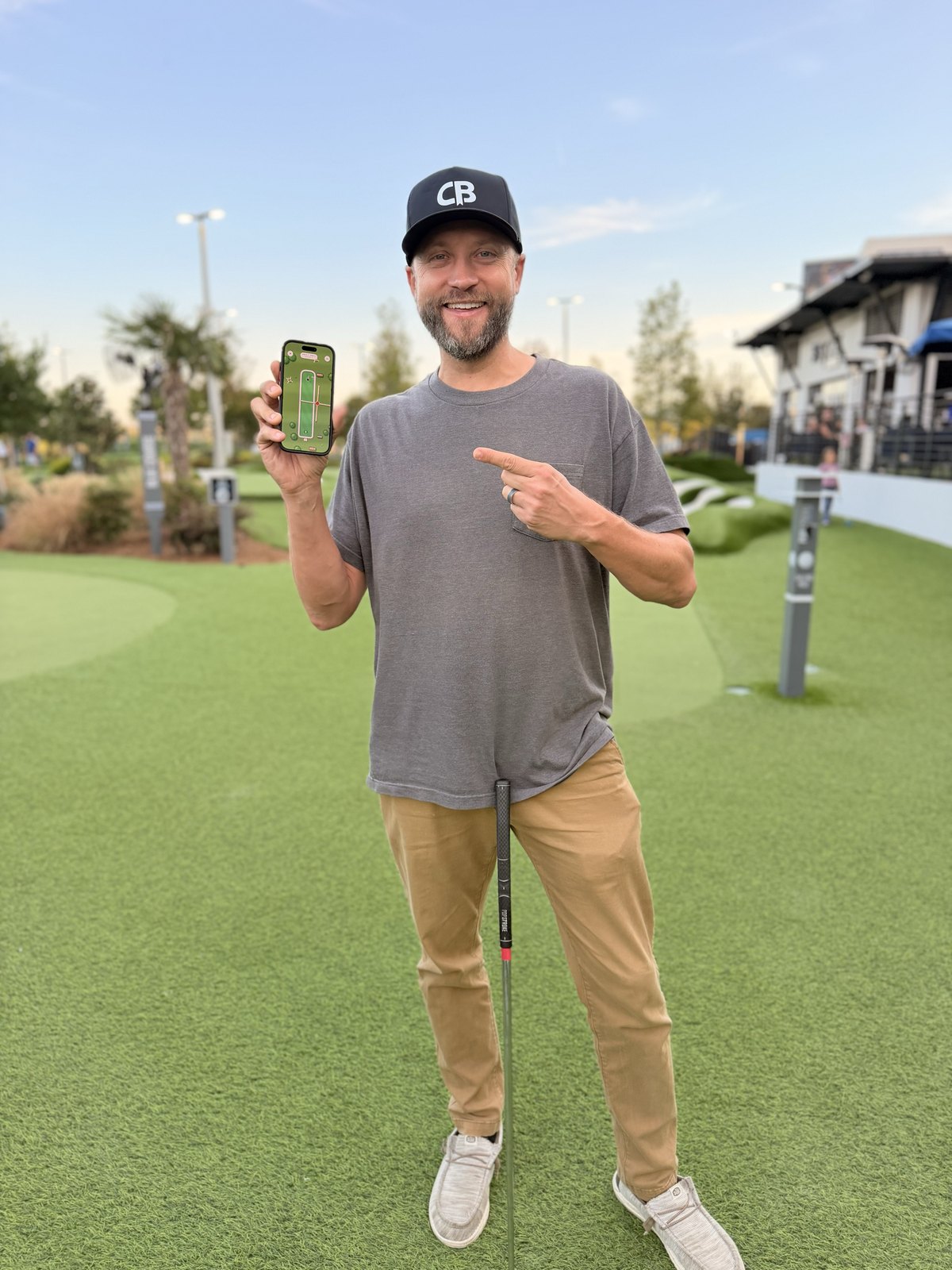 Brian at an outdoor mini golf course at dusk, holding up his iPhone with Greens on the screen and pointing at it
