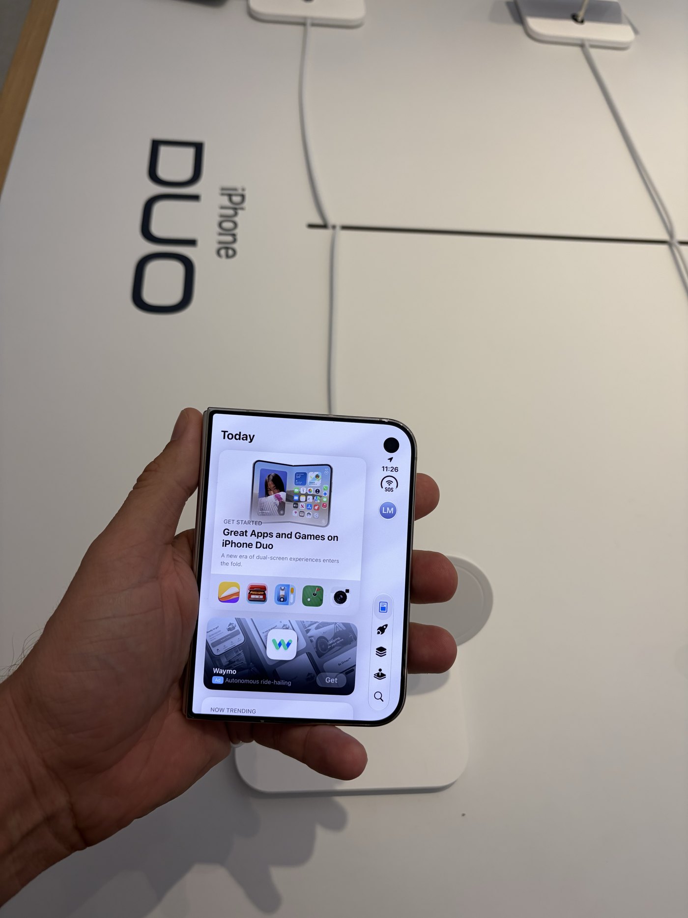 A hand holding an iPhone Duo demo unit at the Apple Store, closed, showing the App Store Today tab with Great Apps and Games on iPhone Duo